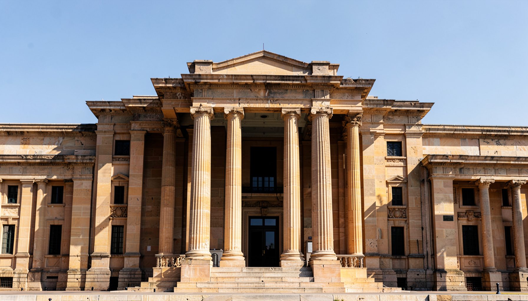 Symmetrical architectural photograph of a stately columned facade in warm sandstone and charcoal tones under a clear sky