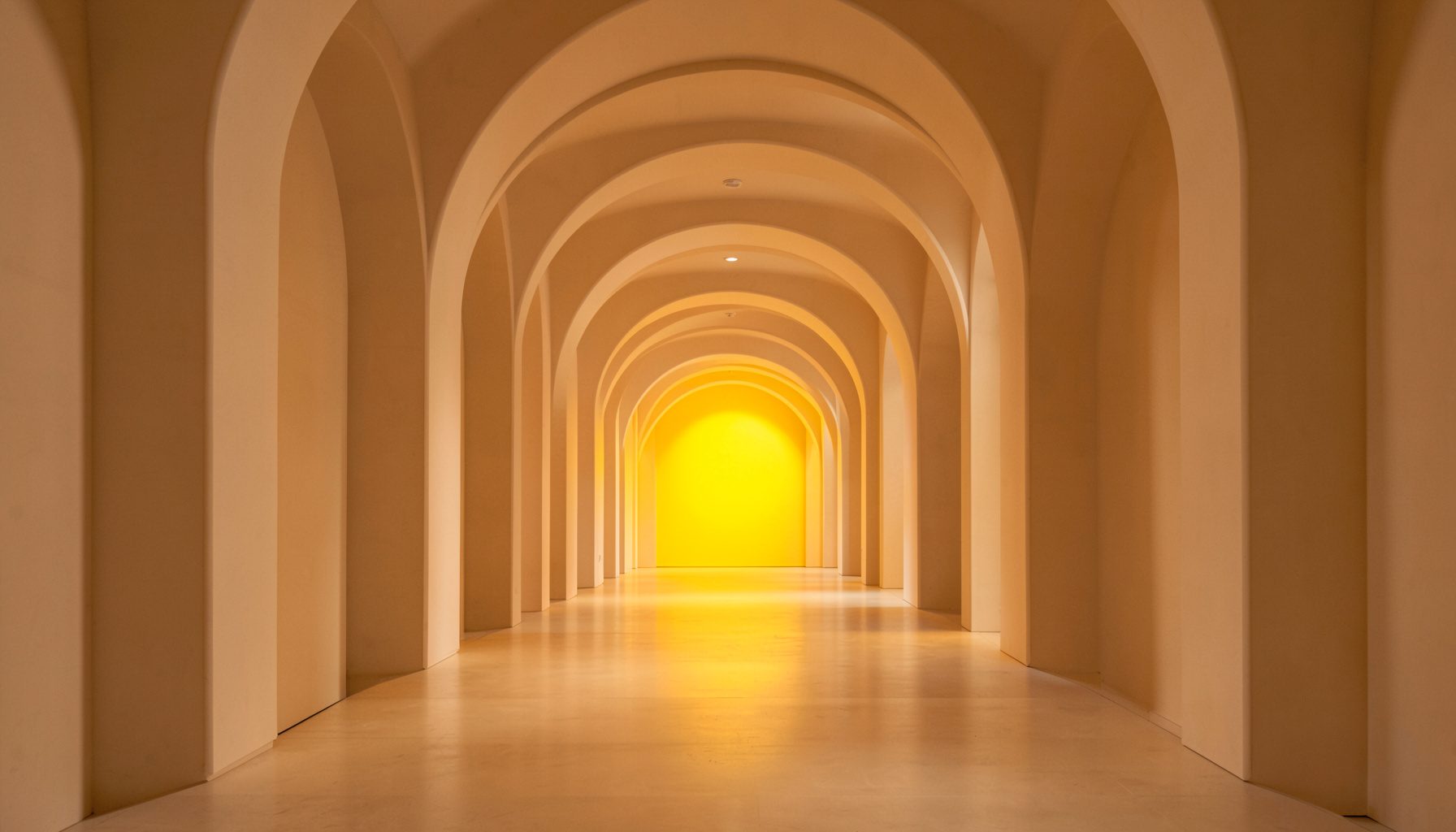 Wide-angle symmetrical photograph of a long interior hallway with repeating arches in warm beige and soft amber light